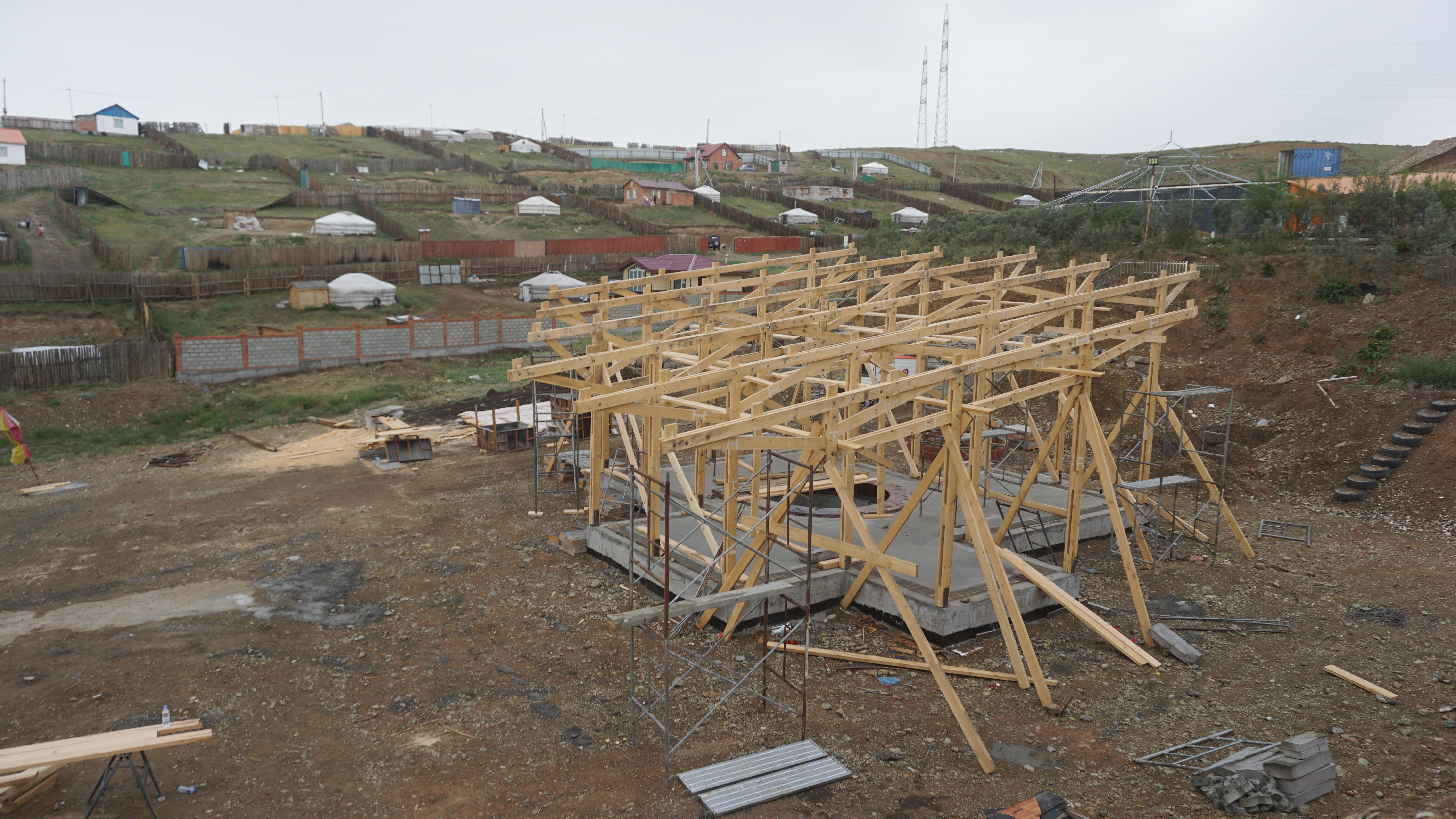 Poured concrete base with a laticced wooden framework roof mid-construction. Individual ger plots can be seen on the hills in the distance. 